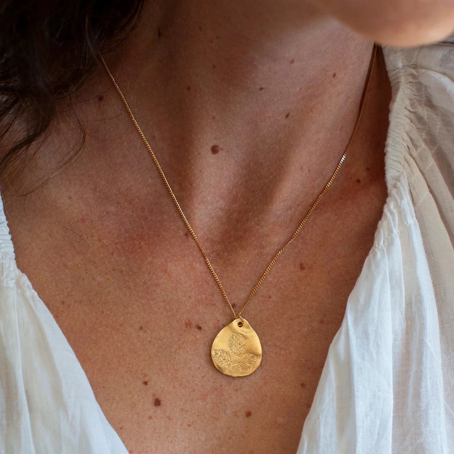 Close-up of a person wearing a gold necklace with a pendant, against a neutral background.