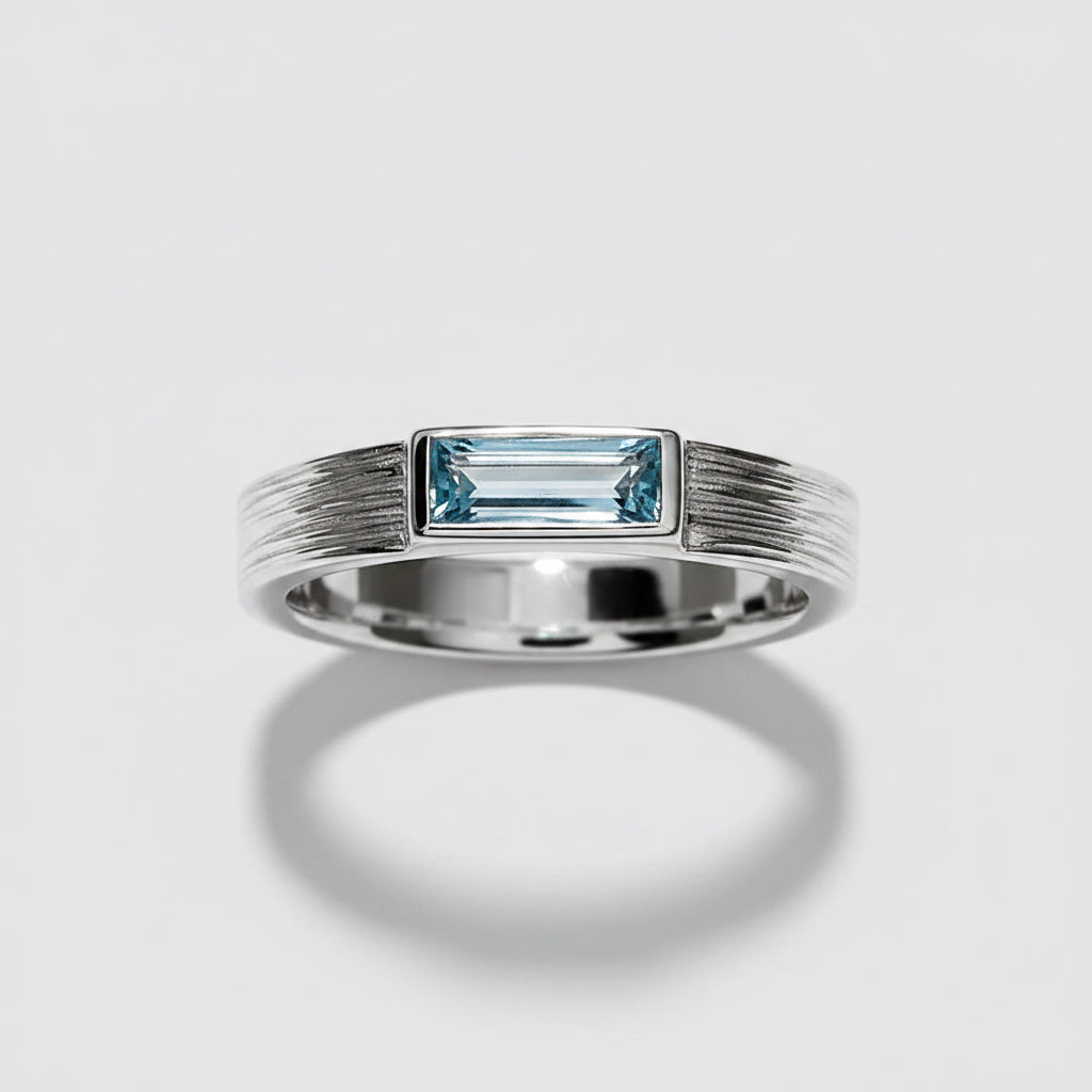 A silver band ring featuring a rectangular baguette-cut light blue gemstone in a bezel setting, with a textured, ribbed design on the shoulders, shown on a white background