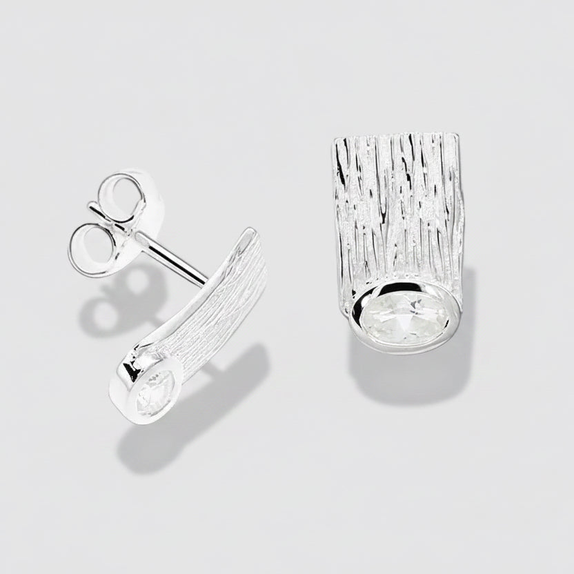 A pair of silver stud earrings on a white background. One earring is shown from the side, displaying a textured, bark-like rectangular face with a clear oval gemstone at the bottom, and the other is shown from the top/front, displaying the post and butterfly clutch back.
