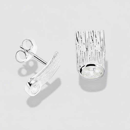 A pair of silver stud earrings on a white background. One earring is shown from the side, displaying a textured, bark-like rectangular face with a clear oval gemstone at the bottom, and the other is shown from the top/front, displaying the post and butterfly clutch back.
